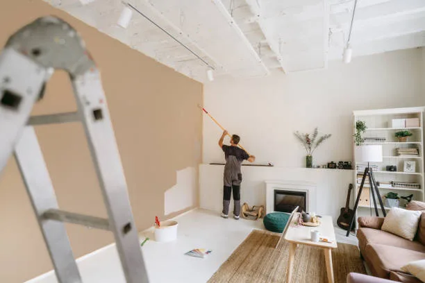 Pintor passando rolo em uma parede de sala, com escada e móveis ao redor